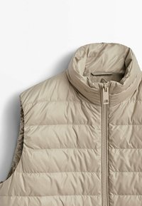 Beżowa ocieplana kamizelka z wysokim kołnierzem, zamkiem błyskawicznym z przodu i pikowanym wzorem. Materiał wydaje się gładki i lekki, o minimalistycznym designie.