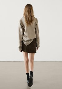 Femme aux longs cheveux marchant, portant une chemise beige à manches longues, une jupe brun foncé, des chaussettes noires et des chaussures noires sur un fond uni.