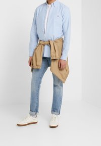 Ljust blå skjorta, beige jacka knuten runt midjan, blå jeans och off-white sneakers med gummisulor. Avslappnad outfit.