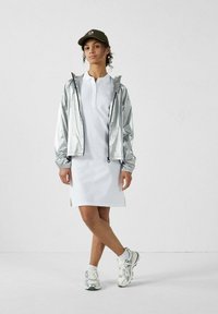 Vrouw draagt een witte polojurk, een metallic zilveren jas, zilveren sneakers, witte sokken en een groene baseballpet, staand tegen een effen witte muur.