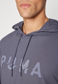 Grauer Kapuzenpullover mit Kordelzug, der ein großes, stilisiertes weißes Logo zeigt. Der Stoff wirkt glatt mit einer weichen Textur.