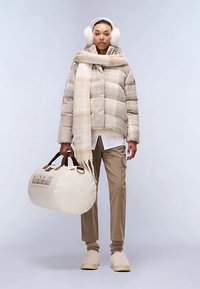 Beež joped pikk talvejope ruudulise mustriga, sobiva salliga, beež püksid ja heledad jalatsid. Käes on ümmargune, kreemikvalge logo ja kotiga.