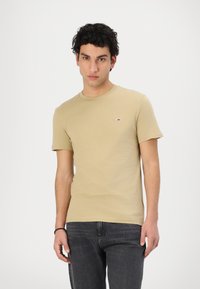 Maglietta a maniche corte color marrone chiaro con collo rotondo, realizzata in cotone, con un piccolo logo sul petto. Indossata con jeans scuri.