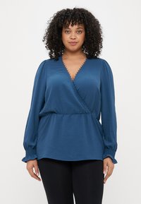 Blouse bleue croisée avec de longues manches bouffantes, présentant une finition texturée et une bordure en dentelle le long de l'encolure et de l'ourlet. Associée à des leggings noirs.
