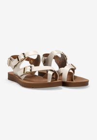 Flache Sandalen mit leichten metallischen Riemen, klobigen Schnallen und einer braunen Gummisohle. Das Design verfügt über eine Zehen-Öse und eine offene Ferse für einfaches An- und Ausziehen.