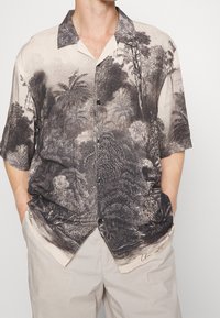 Uomo che indossa una camicia a maniche corte con bottoni, con una stampa dettagliata in bianco e nero di foreste e palme, con le mani nelle tasche di pantaloni beige chiaro.