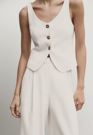 Woman wearing sleeveless white vest with three brown buttons and matching high-waisted wide-leg trousers against plain background.