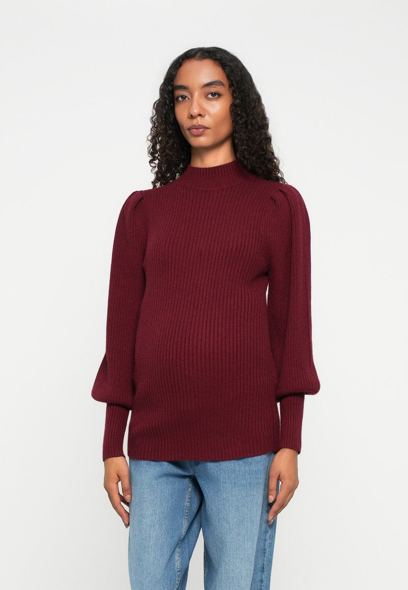 Kvinne med krøllete hår iført en burgunder ribbestrikket genser med puffede ermer og blå jeans, stående mot en enkel hvit bakgrunn.