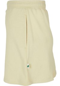 Pantalones cortos de algodón de color amarillo claro con cinturilla elástica, bolsillos laterales y una pequeña etiqueta con un logo de hoja verde en el dobladillo. Textura suave.