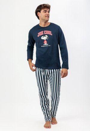 Joven con camiseta azul marino de manga larga "Joe Cool" de Snoopy y pantalones blancos con rayas verticales azul marino, de pie descalzo sobre un fondo blanco.