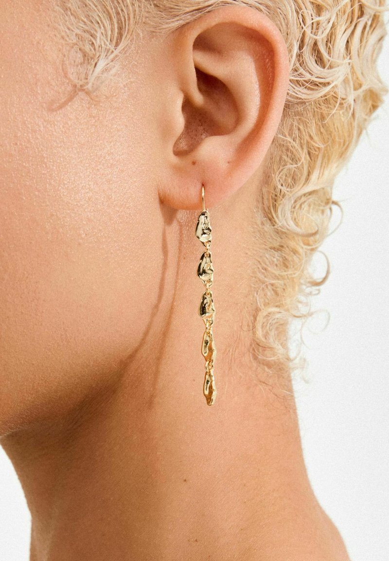 Gold dangle earrings feature a textured design with irregular shapes, hanging below the earlobe, showcasing reflective surfaces and a shiny finish.