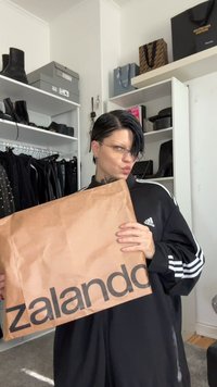Osoba s krátkymi tmavými vlasmi, oblečená v čiernom oblečení Adidas, drží veľký hnedý balík Zalando v miestnosti so poličkami na obuv a tašky.