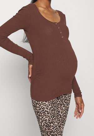 Femme enceinte portant un haut côtelé marron à manches longues et un legging imprimé léopard, avec une main sur le bas du dos.