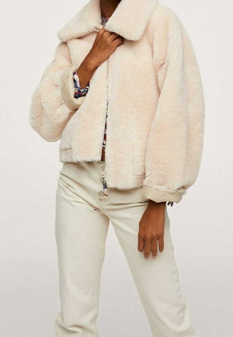 Personne portant une veste crème, duveteuse avec de larges manches et un pantalon beige clair, debout devant un fond uni.