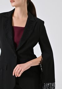 Blazer nero con colletto in pizzo testurizzato e dettagli in pizzo sulle maniche, abbinato a una maglia bordeaux scura. Tessuto morbido e taglio sartoriale.