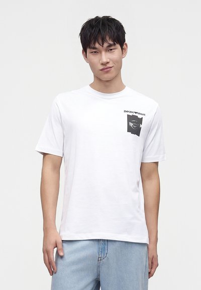 Νέος άνδρας που φοράει ένα λευκό t-shirt Emporio Armani με μικρό μαύρο γραφικό στο αριστερό στήθος και ανοιχτόχρωμο τζιν, στέκεται μπροστά σε μία απλή φόντο.