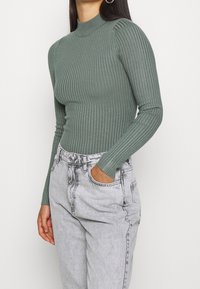 Vrouw in een groene ribgebreide langetermijn coltrui en lichtgrijze hoge taillejeans met één hand in de zak.