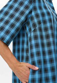 Mano che riposa all'interno della tasca di una camicia a maniche corte con un motivo a quadri blu e nero, che mostra la texture del tessuto e i dettagli della cucitura.