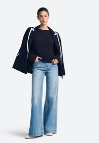 Granatowy płaszcz z białymi akcentami, czarny sweter z prążkowanym wzorem oraz jasnoniebieskie dzwonowate dżinsy. Modelka ma na sobie białe buty na szpic, ręce w kieszeniach.