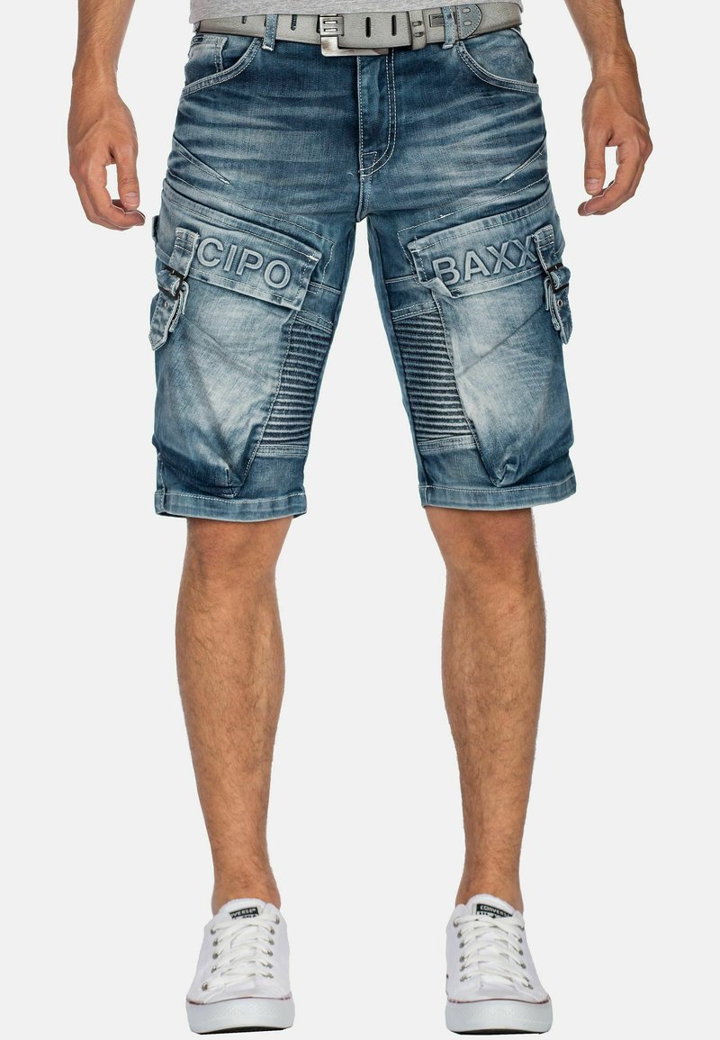Pantaloni cargo in denim blu sbiadito con molteplici tasche, dettagli testurizzati e cuciture bianche. Abbinati a una cintura grigia e sneakers bianche.