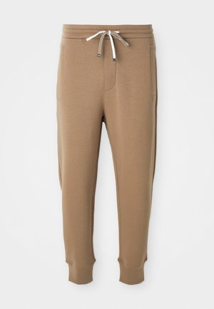 Pantalons joggers beige clair avec une taille élastique, des cordons de serrage blancs et marron, des poches latérales et des chevilles côtelées sur fond blanc.