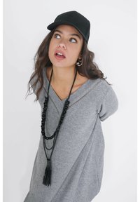 Grijze gebreide trui met een V-hals, zwarte kraalhalsketting met een kwastje en een zwarte baseballcap. Soepele textuur en een ontspannen pasvorm.