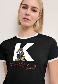 Schwarzes T-Shirt mit kurzen Ärmeln, das ein großes weißes "K", eine Cartoon-Ente in Flugzeugpilotenkleidung und darunter roten Schriftzug zeigt. Gerippter weißer Ausschnitt.