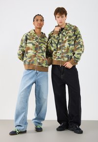 Sudaderas con capucha de camuflaje en verde, marrón y beige, combinadas con jeans ligeros y holgados en azul claro y negro. Zapatillas con cordones verdes.