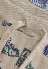 Beige Baumwollstoff mit blauen und grünen Traktor-Designs; verfügt über einen gerippten Taillenbund und einen geflochtenen Kordelzug mit beigen Ösen.