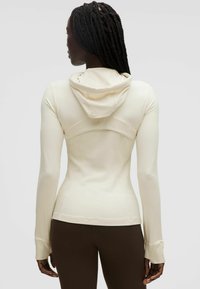 Sudadera ajustada de un color beige claro con cuello alto y capucha, que cuenta con mangas largas y detalles de costura a lo largo de las costuras.