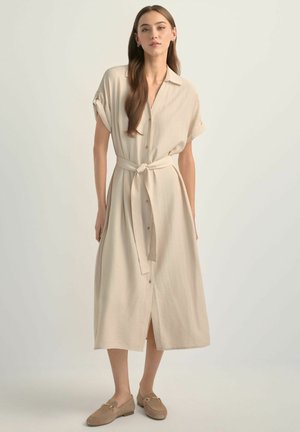 Kobieta o długich brązowych włosach, ubrana w beżową, zapinaną na guziki sukienkę midi z paskiem w talii i pasujące mokasyny, stojąca na jednolitym jasnym tle.