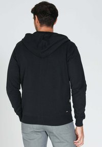Sweatshirt à capuche noir avec manches longues, bords-côtes aux poignets et à l'ourlet, et une petite étiquette logo en bas à gauche. Matériau en mélange de coton doux.