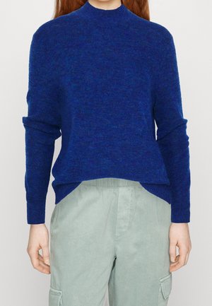 Personne portant un pull bleu à manches longues et un pantalon cargo gris clair, debout devant un fond uni.