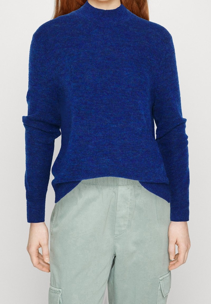 Personne portant un pull bleu à manches longues et un pantalon cargo gris clair, debout devant un fond uni.