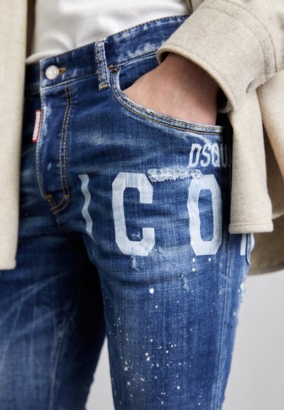 Blaue, verwaschene Jeans mit einem auffälligen weißen Druck und Spritzereffekten. Der Stoff hat eine ausgebleichte Textur und fransige Details.