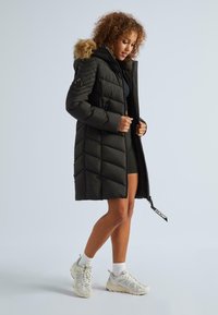 Sieviete pielāgo garu melnu quilt-coat ar kažokādu, ģērbusies melnā kleitā, baltās zeķēs un baltās sporta apavos, stāvot pret gaismas fona.