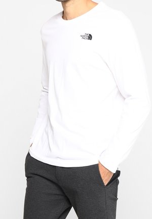 Mężczyzna w białej koszulce z długim rękawem z logo The North Face na klatce piersiowej i ciemnoszarych spodniach, ręce w kieszeniach.