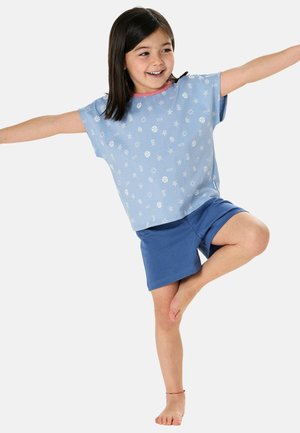 Young girl balancing on one leg with arms extended, wearing blue shorts and a light blue patterned t-shirt, smiling and looking sideways.