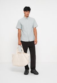 Light blue short-sleeve button-up shirt, dark pants, holding a cream tote bag. Features a chest pocket and soft, textured fabric.