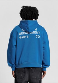 Blauwe hoodie met een grote witte grafiek op de achterkant, met de tekst "CREATIVE DEPARTMENT 2018 CD". Losse pasvorm, zachte textuur en een kangoeroezak.