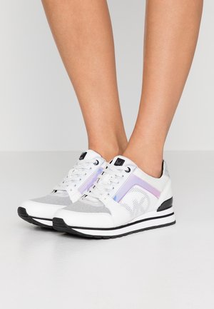 Hvide og grå snøresko med iriserende sidepaneler båret på bare fødder, vist mod en ensfarvet lys baggrund.