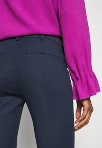 Fialová bluzka s texturou a volány na rukávech spárovaná s tmavě námořnickými kalhotami na míru s zadními kapsami. Záběr zblízka na ruku na pásu.