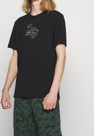 Person trägt ein schwarzes T-Shirt mit farbigem "ZIM"-Text und grünen Camouflage-Shorts, steht vor einem einfarbigen hellen Hintergrund.