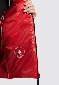 Rød pufferjakke med inderside, der har en skinnende tekstur, trykt logo "KHUJO GENUINE" med et ahornblad og sorte lynlåsdetailler.