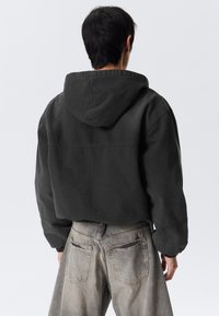Persoon met kort donker haar, gekleed in een cropped zwart hoodie-jack en vervaagde grijze jeans, staande met de rug naar de camera tegen een effen achtergrond.