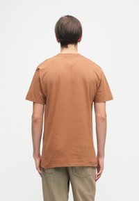 Καφέ βαμβακερό T-shirt με κοντά μανίκια, στρογγυλή λαιμόκοψη και άνετη εφαρμογή. Η ορατή υφή είναι λεία, με ένα απλό, ακατέργαστο σχέδιο.