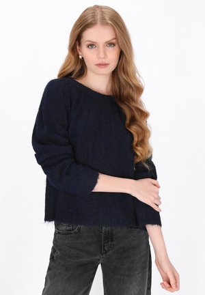 Jonge vrouw met lang golvend blond haar, pareloorbellen, een donkerblauwe zachte trui en een zwarte spijkerbroek, tegen een witte achtergrond.