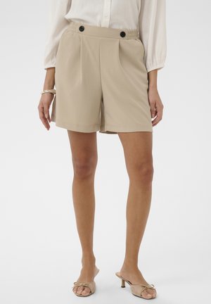 Žena v béžových vysokých šortkách s čiernymi gombíkmi, bielou blúzkou, béžovými sandálmi na podpätku a zlatým náramkom na ľavej ruke.