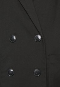 Náhled na přední část černého dvouvrstvého kabátu se čtyřmi černými knoflíky a viditelným okrajem kapsy na textuře látky.