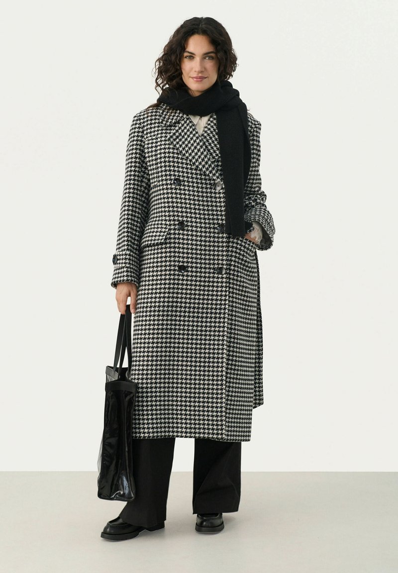 Black and white houndstooth double-breasted coat, textured wool fabric, paired with a long black scarf and sleek black bag. Wide-leg pants and low shoes.